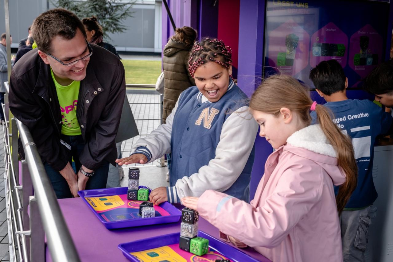 A volunteer wearing a bright green SPARK™ T-shirt smiles as two students stack learning cubes on activity trays inside the Curiosity Cube™ during a hands-on STEM activity.