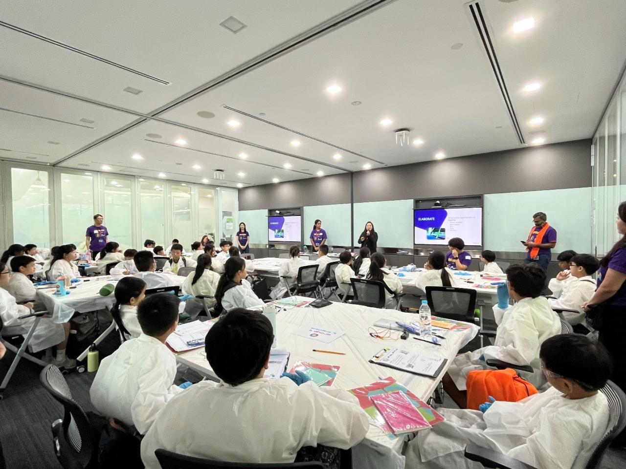 Students wearing white lab coats sit around tables as volunteers in purple SPARK™ shirts lead a Curiosity Labs™ workshop.