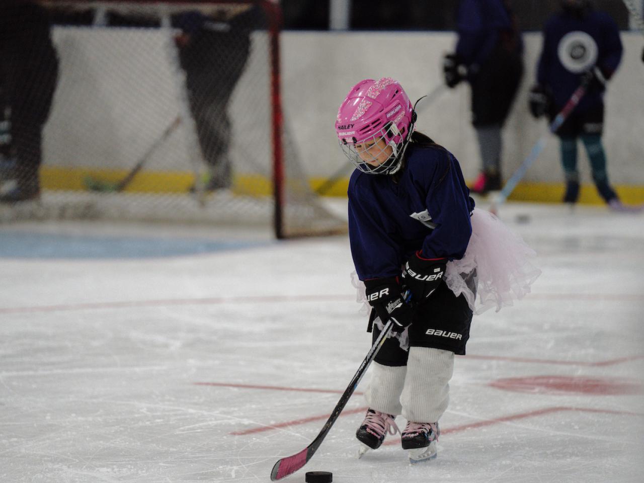 Participant in full hockey gear skates on the ice while practicing puck control during LA Kings Girls Camp. A pink helmet and matching hockey stick stand out against the rink background.