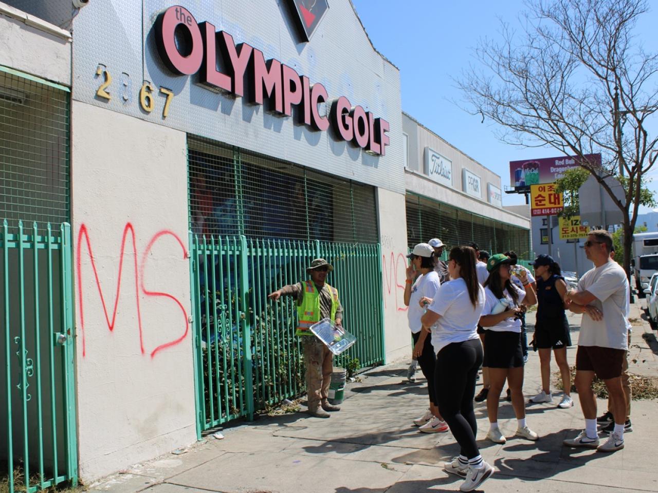 Community volunteers gather on a sidewalk as a KYCC cleanup leader provides instructions in front of a building marked with graffiti.