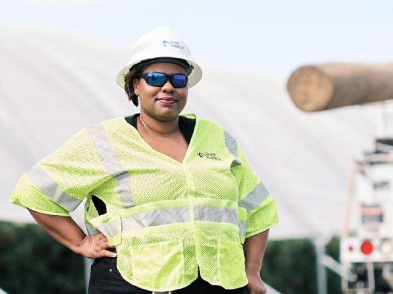LaQuitta Ghent next to large equipment, wearing high-vis vest at hard hat. One hand on a hip.