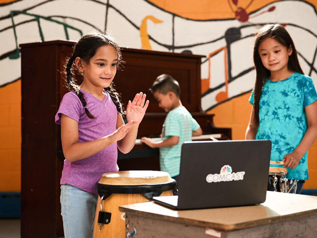 children standing in front of a laptop, one behind them playing on a piano. Wall behind them with a painted mural of floating music notation.