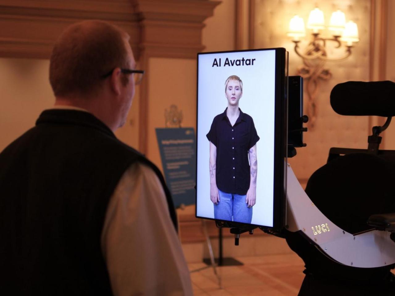 Erin Taylor appearing as an avatar on a small screen, another looks at it.