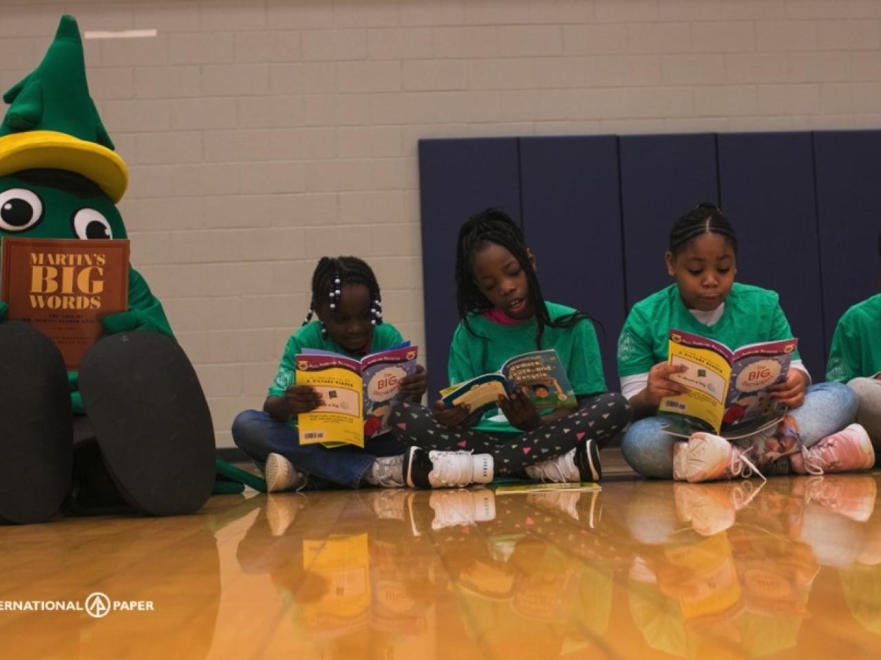 Children sitting in a line on a gym floor, a tree mascot next to them, all reading books.