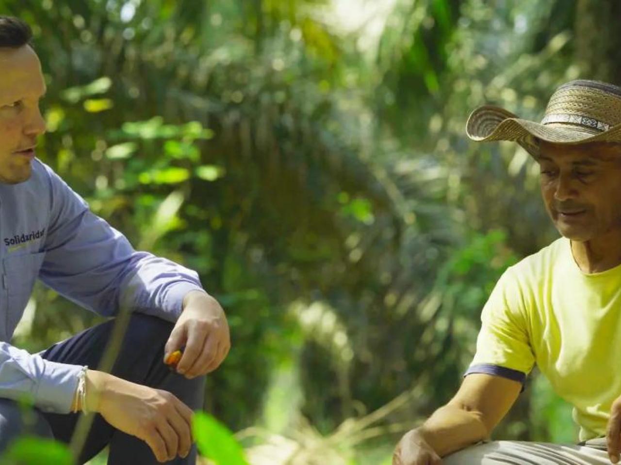 Two people crouched, talking together, in a jungle setting.