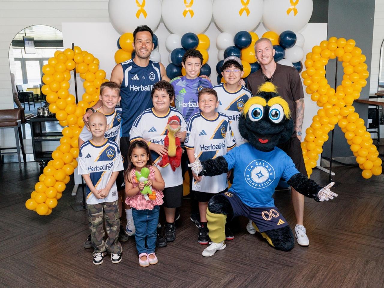 Youth participants, LA Galaxy players, and the LA Galaxy Foundation mascot Cozmo pose together in front of gold ribbon balloon displays during a community engagement event.
