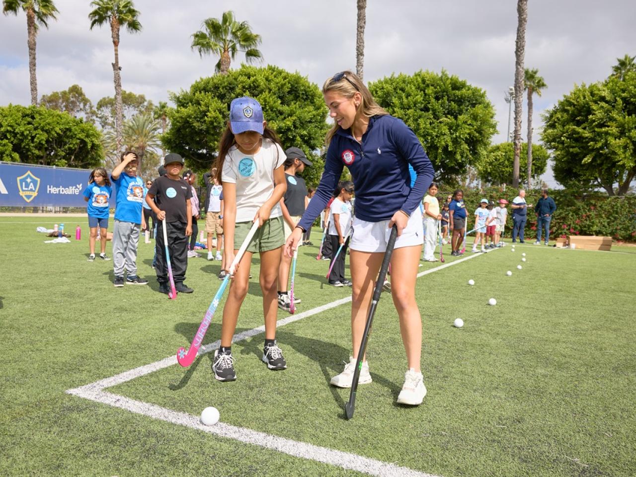 Ashley Sessa demonstrates field hockey stick positioning to a student as other students practice with sticks and balls on the field.