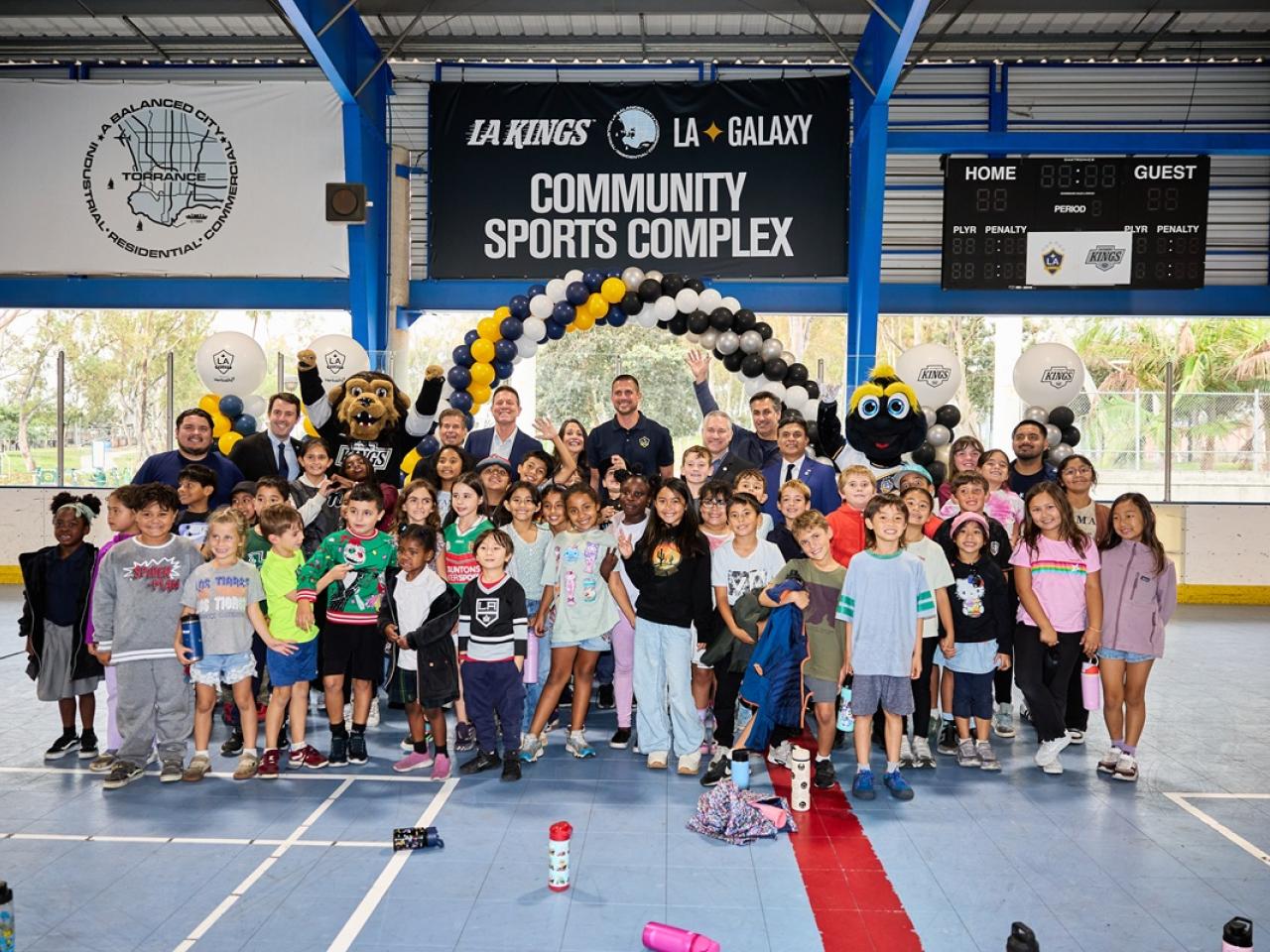 A large group of children and adults pose with the LA Kings and LA Galaxy mascots beneath signage for the LA Kings and LA Galaxy community sports complex in Torrance.
