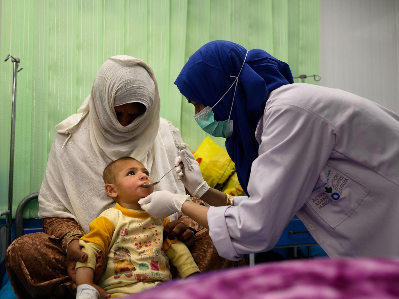 An ACF nurse leans in to give a spoonful of liquid medicine to a toddler. 