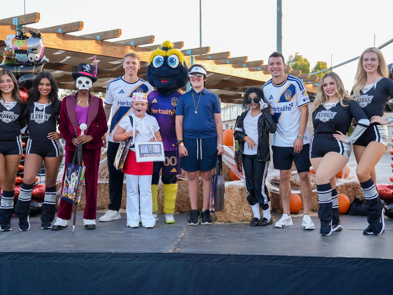 LA Kings Ice Crew members, LA Galaxy representatives, a costumed mascot and children in Halloween costumes pose together on a decorated outdoor stage.