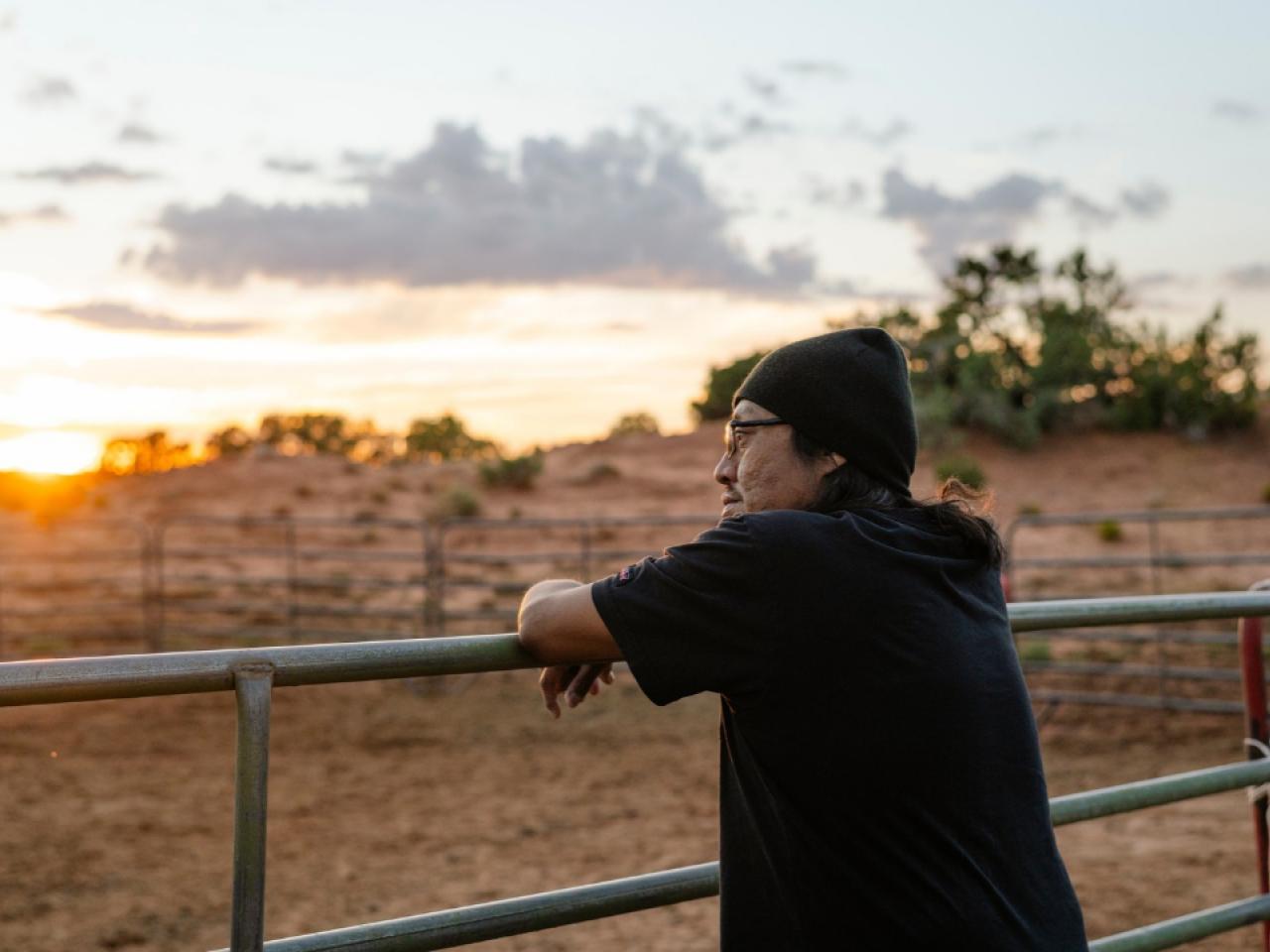 Nobert, a DaVita home dialysis patient, leans against a metal farm fence while he looks out at a sunset on Navajo Nation.