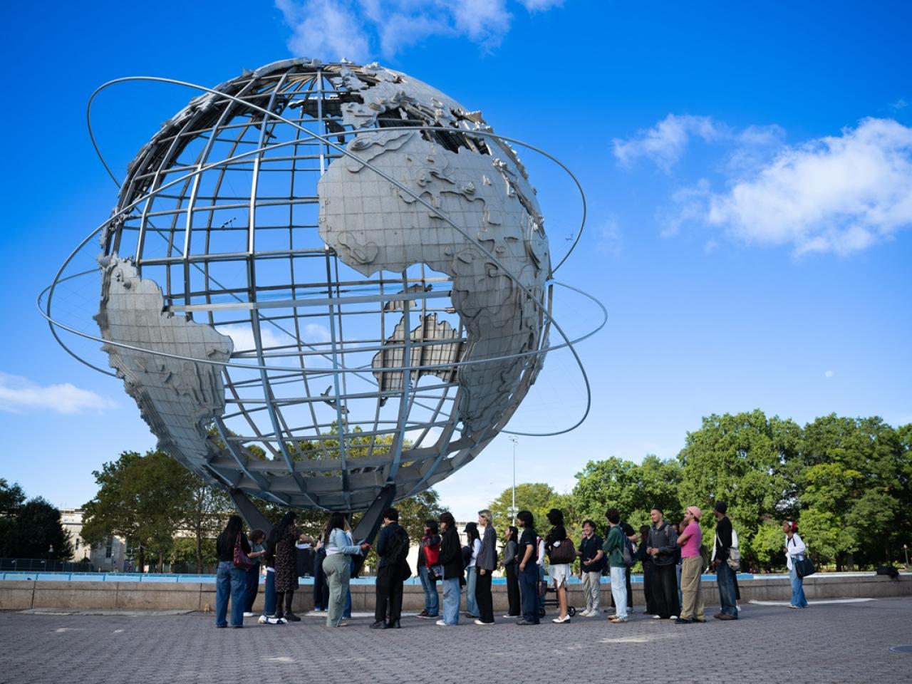 Group gathers outdoors beneath the Unisphere at Flushing Meadows Corona Park under a bright blue sky.