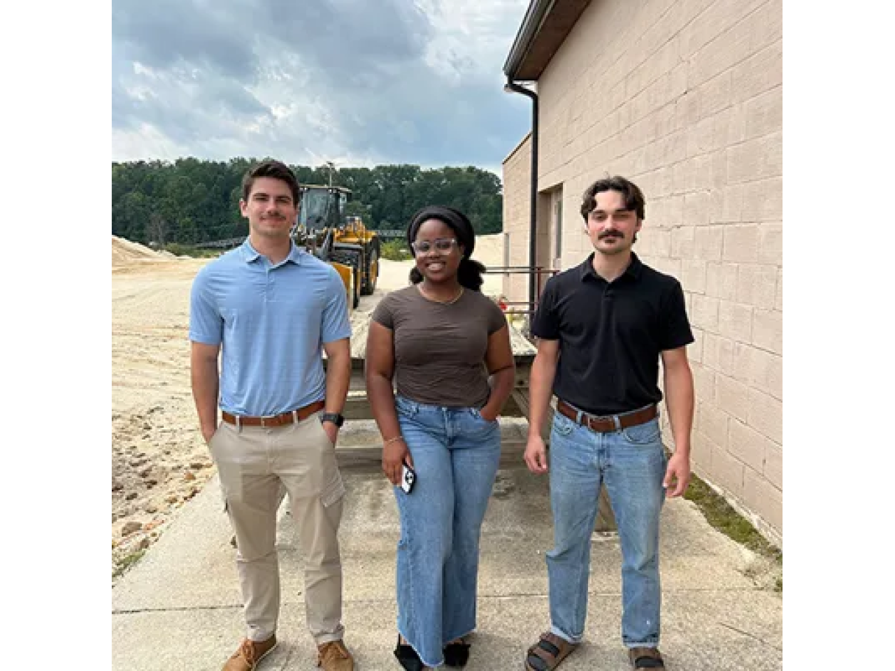 The three trainees standing together next to a building, with a tractor and trees in the background