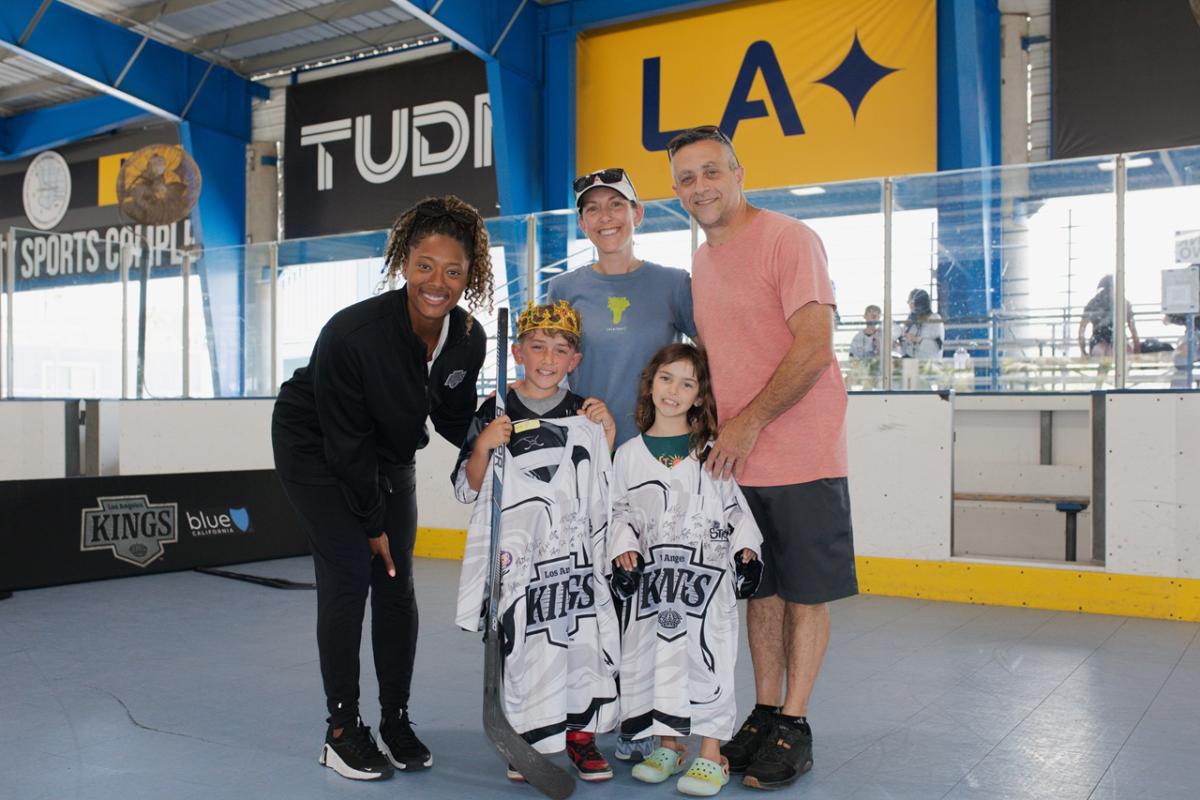Two youth participants stand with signed LA Kings jerseys alongside family members and LA Kings Blake Bolden during a ball hockey event at an indoor sports facility.