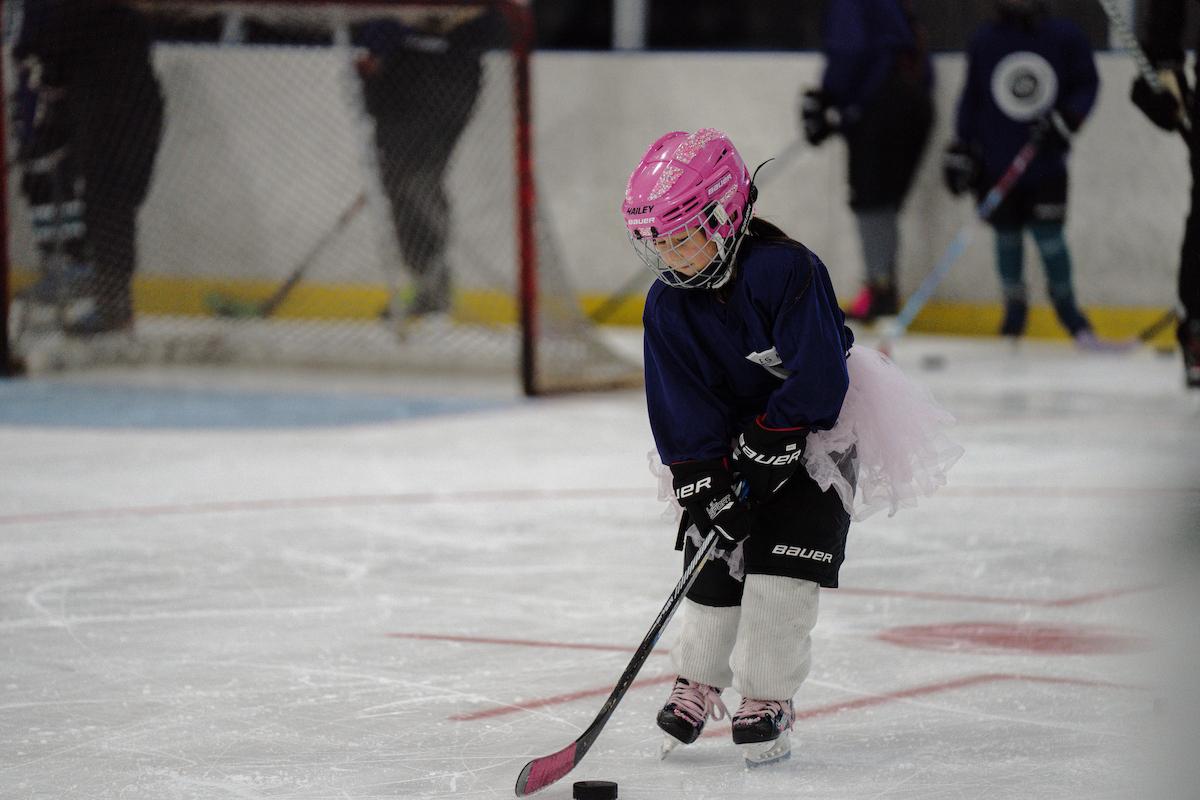 Participant in full hockey gear skates on the ice while practicing puck control during LA Kings Girls Camp. A pink helmet and matching hockey stick stand out against the rink background.