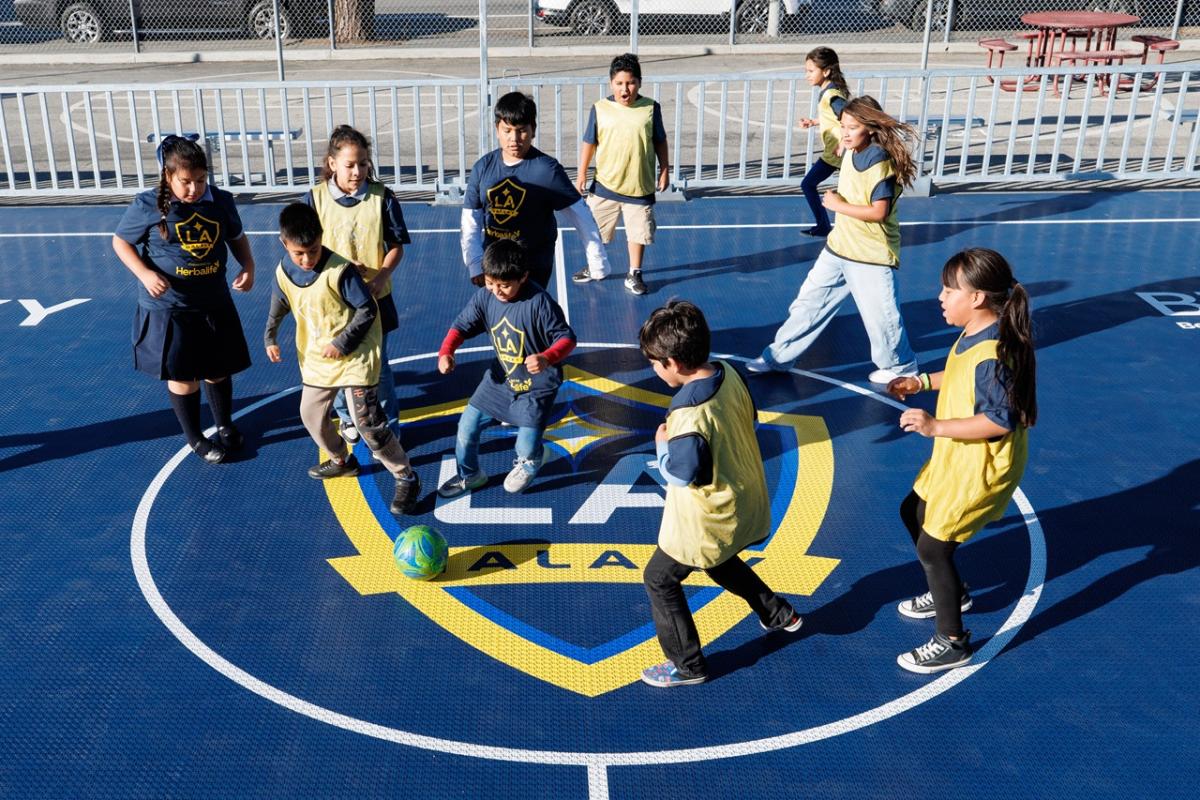 Children wearing LA Galaxy jerseys and pinnies play soccer around a ball on a branded mini-pitch court.