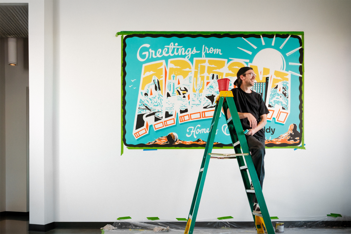 Artist on ladder in front of mural which reads "Greetings from Arizona"