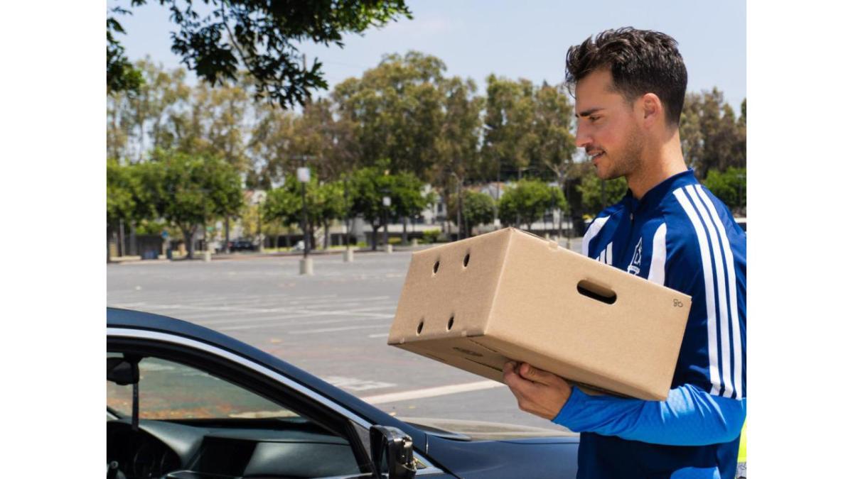LA Goalkeeper JT Marcinkowski in a blue and white jacket holds a food donation box next to a dark car in a parking lot.