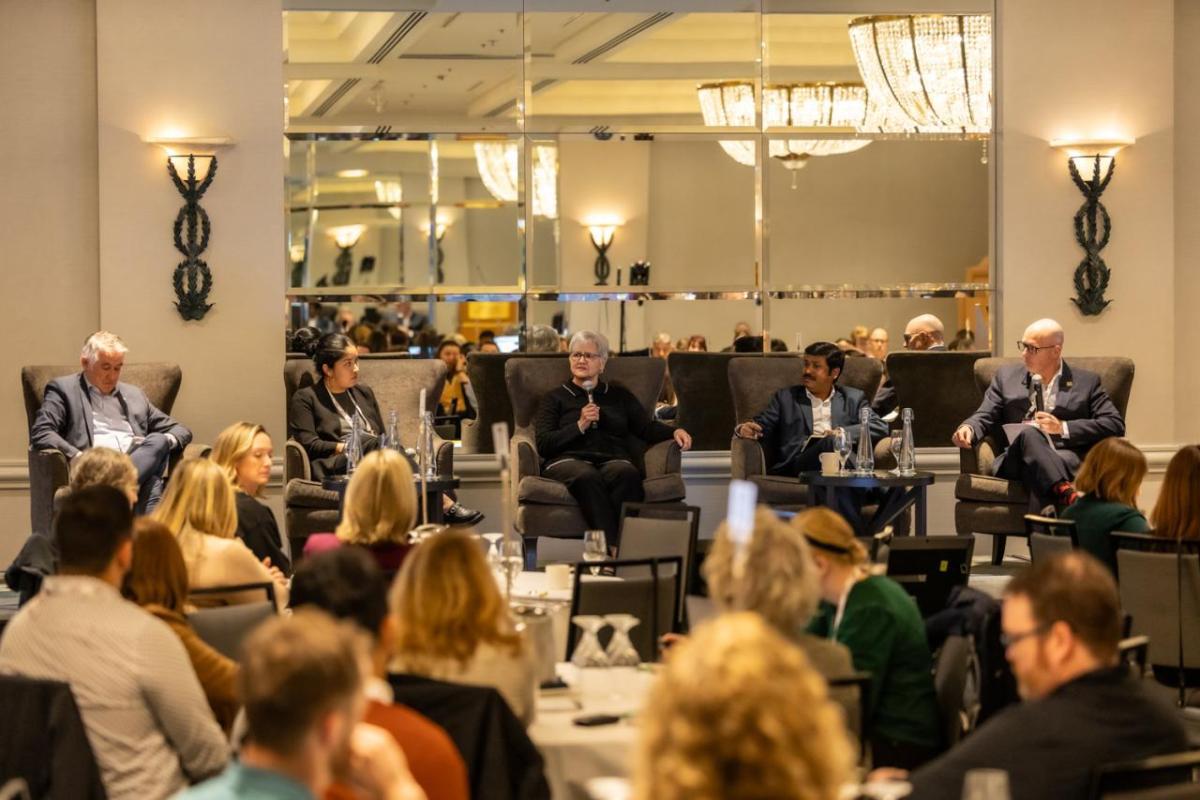 A panel of five people speaking and answering questions at a conference event, seated on stage in front of an audience.