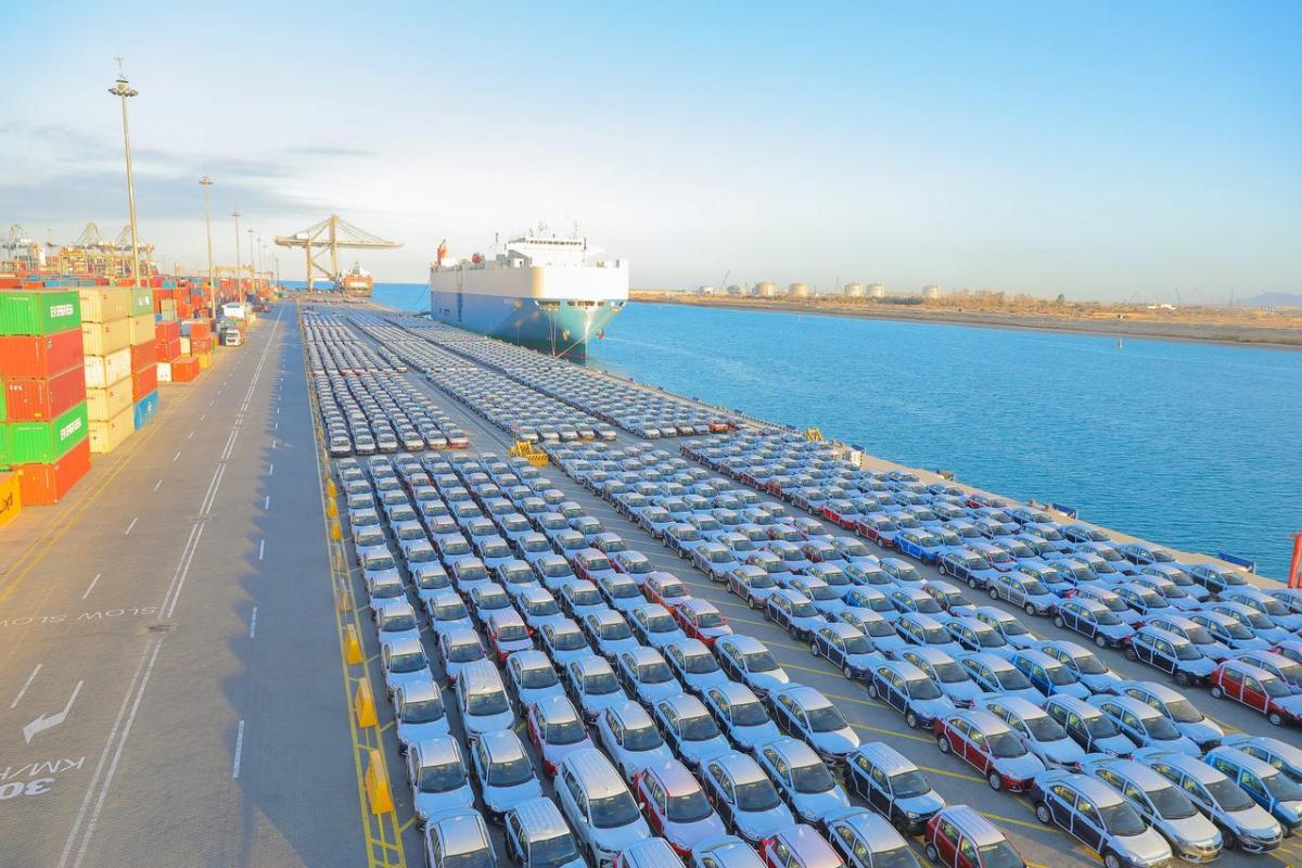 Rows of finished vehicles staged at a port terminal alongside a cargo vessel, illustrating large-scale automotive imports and exports supported by integrated logistics operations.
