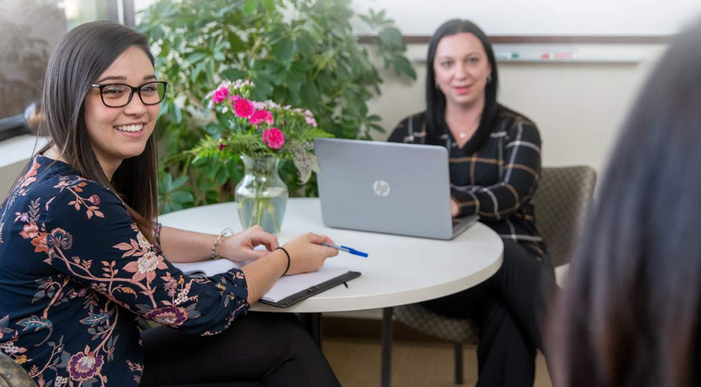 Two people sitting at a round table, both turned looking off screen. One has an open laptop.