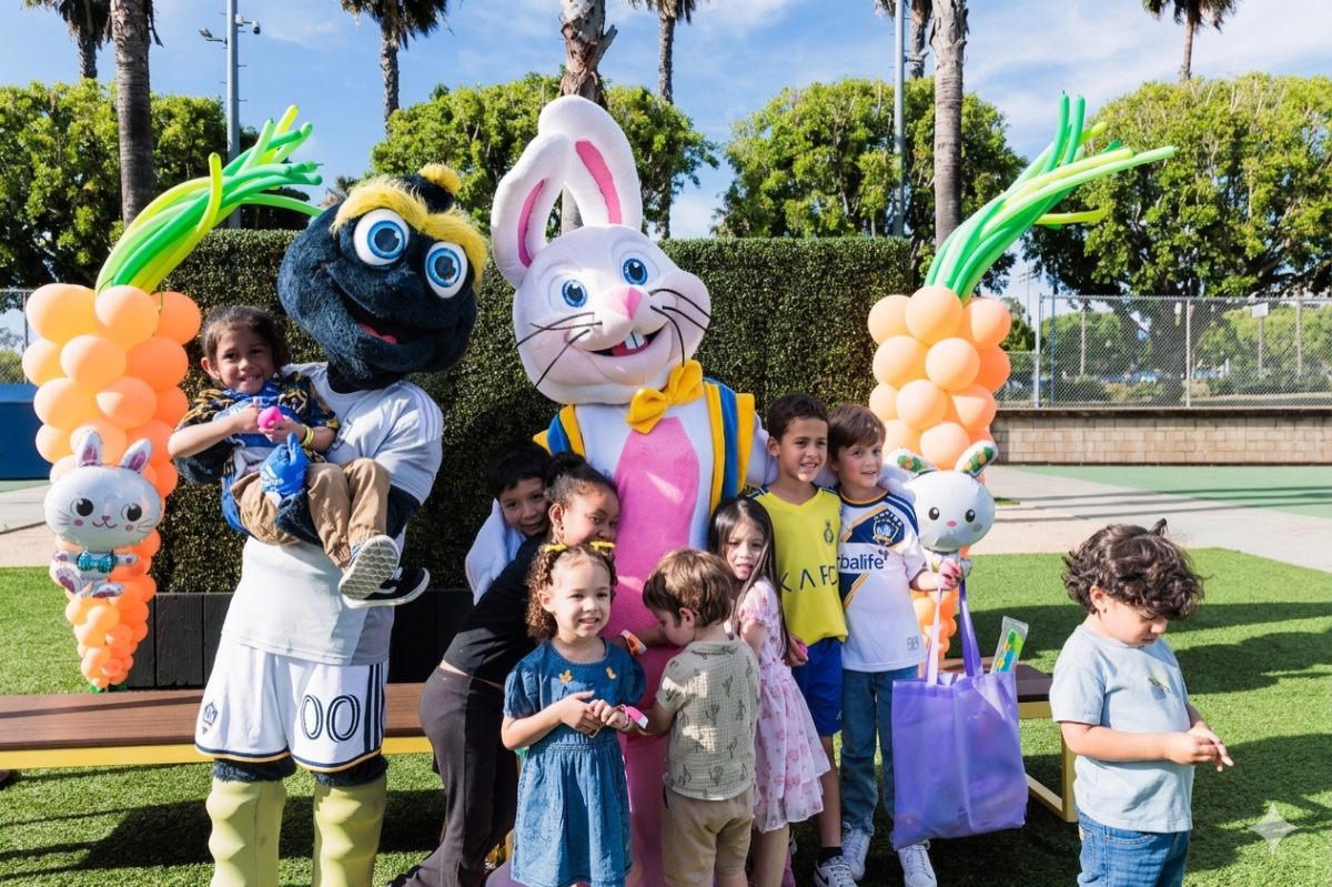Children gather with two mascots on a turf field, holding gift bags and standing beneath balloon decorations.