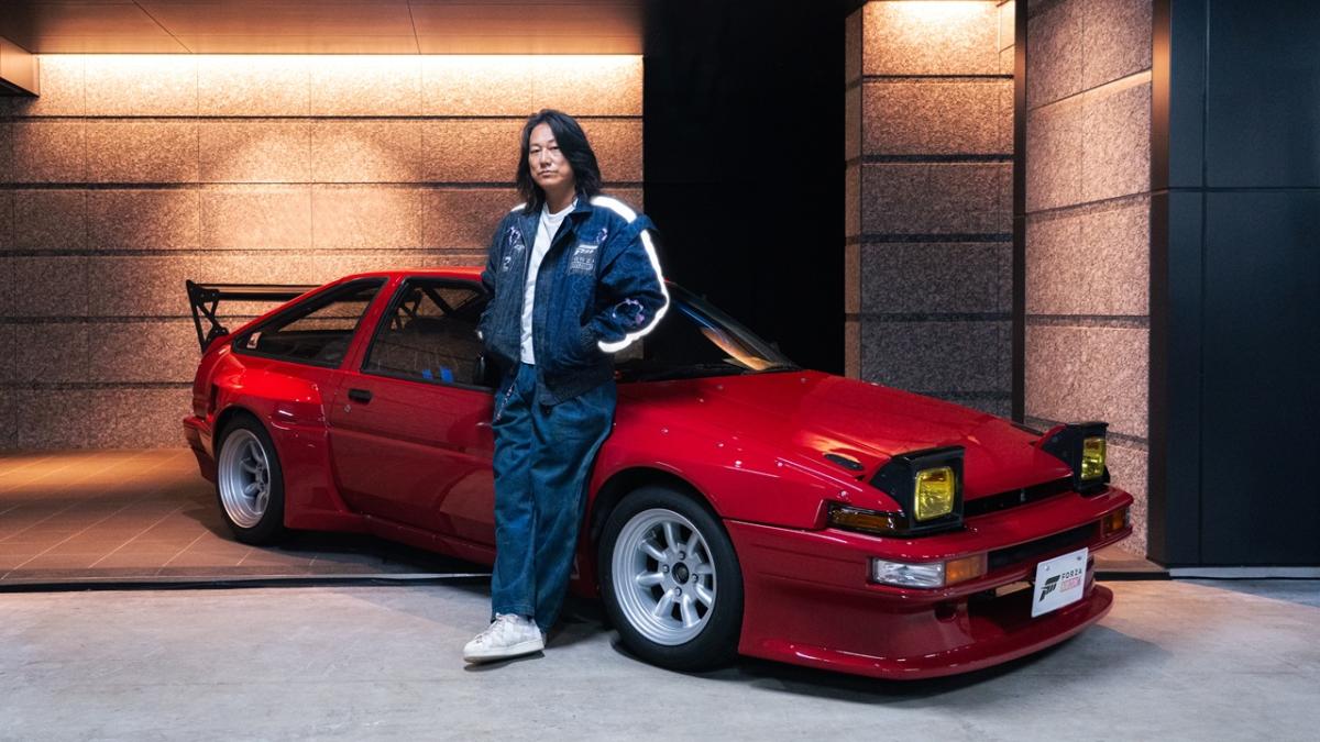 Sung Kang leaning against a red sports car with pop-up headlights in a modern garage, wearing a dark jacket and loose-fit pants.