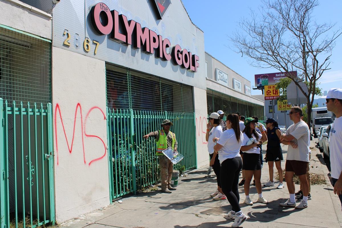 Community volunteers gather on a sidewalk as a KYCC cleanup leader provides instructions in front of a building marked with graffiti.
