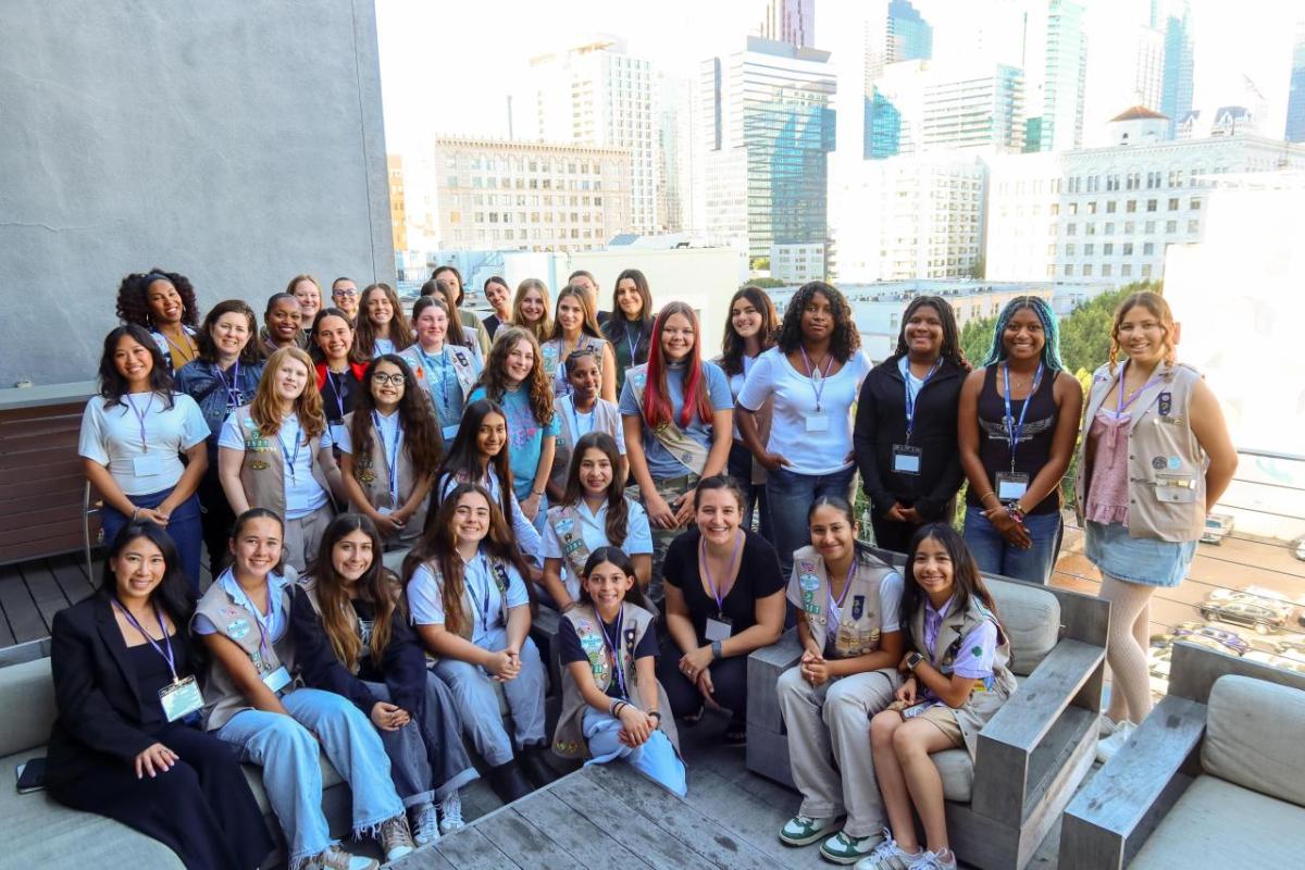 A large group of participants gathers on an outdoor terrace with city buildings in the background, posing together during a group activity.