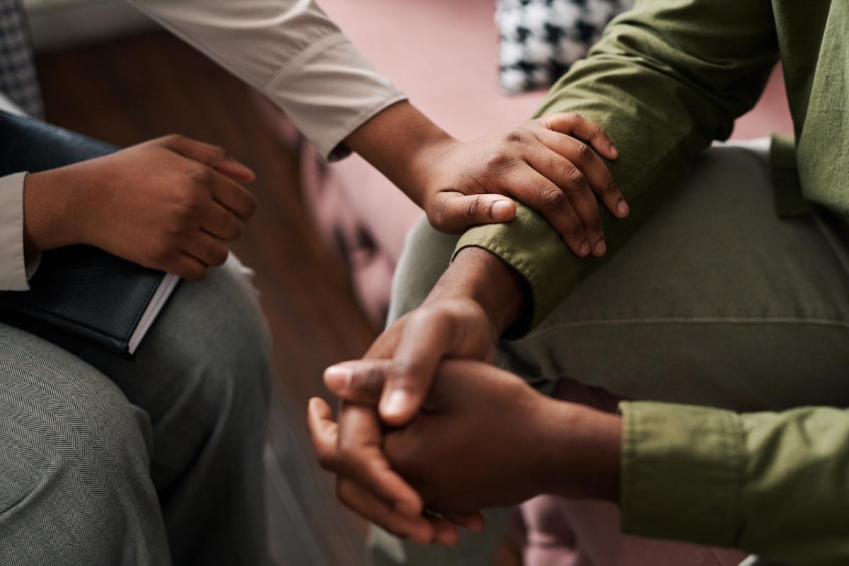 Two people seated. One with folded hands and the other placing his arm in a reassuring manner on the others arm.