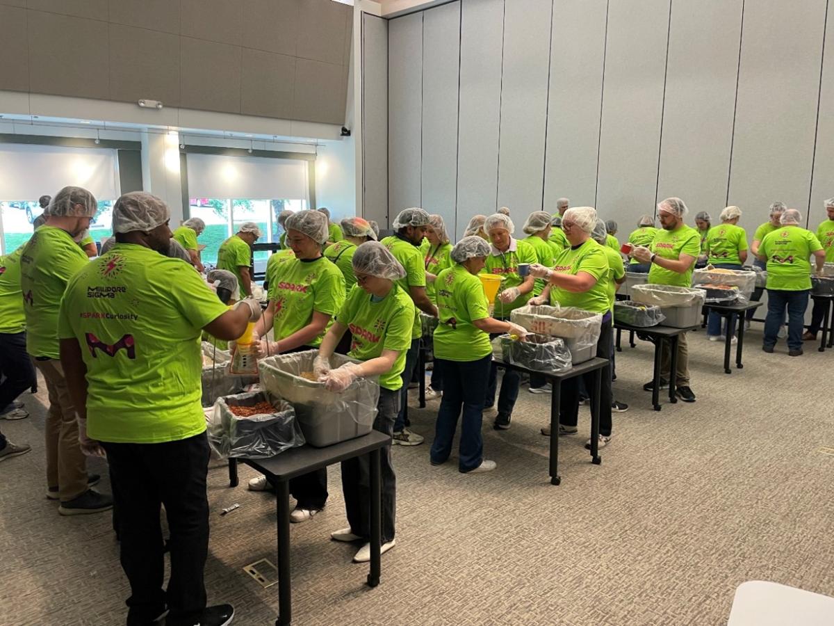 Volunteers wearing bright green SPARK™ t-shirts, hairnets, and gloves assemble meal kits.