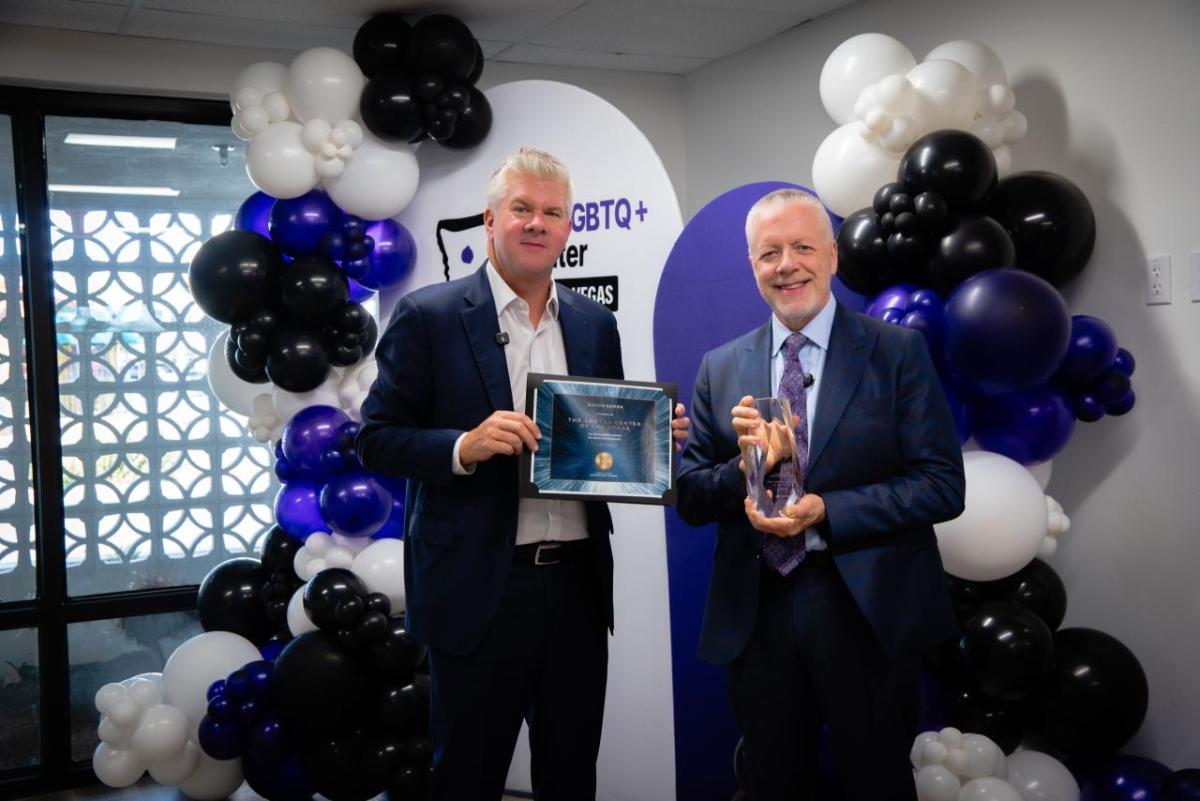 Two men standing in front of balloons. One is holding a framed certificate and one is holding an award.