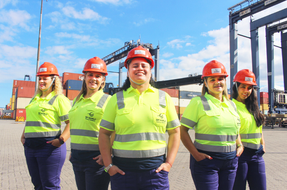 Five women wearing safety gear stand in front of container cranes and stacked shipping containers at DP World’s Santos terminal in Brazil.