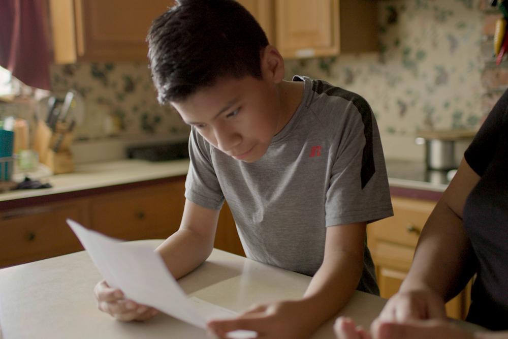 Young boy reading from a piece of paper at a kitchen table.