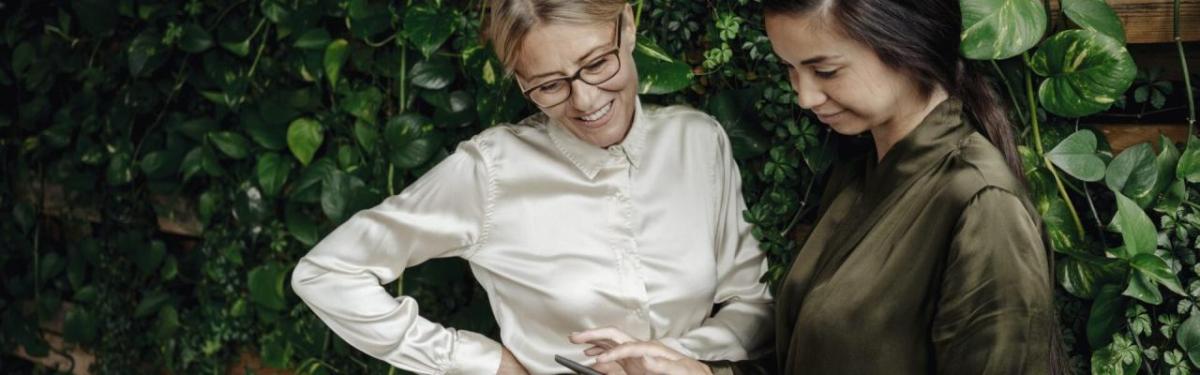 Two people looking at something together and smiling, in front of greenery