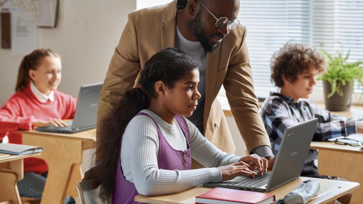 A teacher helping a student in a classroom, using a laptop