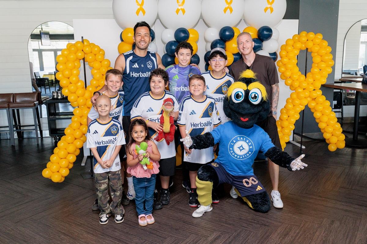 Youth participants, LA Galaxy players, and the LA Galaxy Foundation mascot Cozmo pose together in front of gold ribbon balloon displays during a community engagement event.
