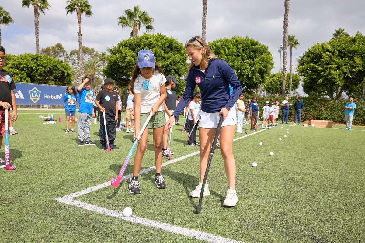 Ashley Sessa demonstrates field hockey stick positioning to a student as other students practice with sticks and balls on the field.