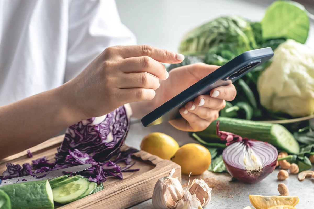 A person with a smart phone and fruits and vegetables on a counter.