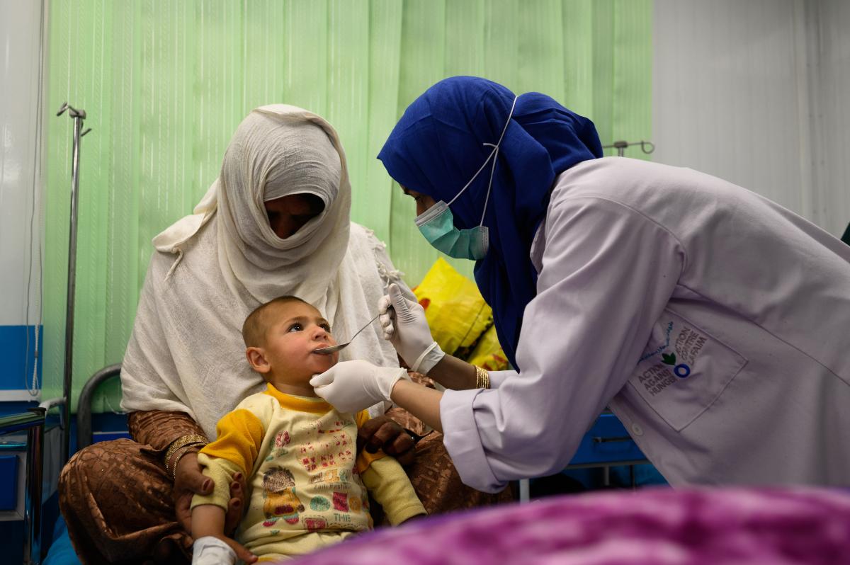 An ACF nurse leans in to give a spoonful of liquid medicine to a toddler. 