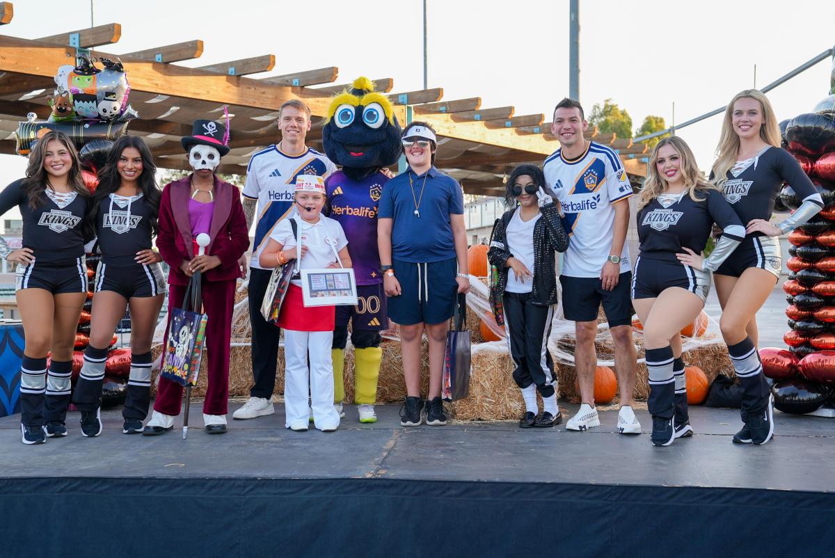 LA Kings Ice Crew members, LA Galaxy representatives, a costumed mascot and children in Halloween costumes pose together on a decorated outdoor stage.
