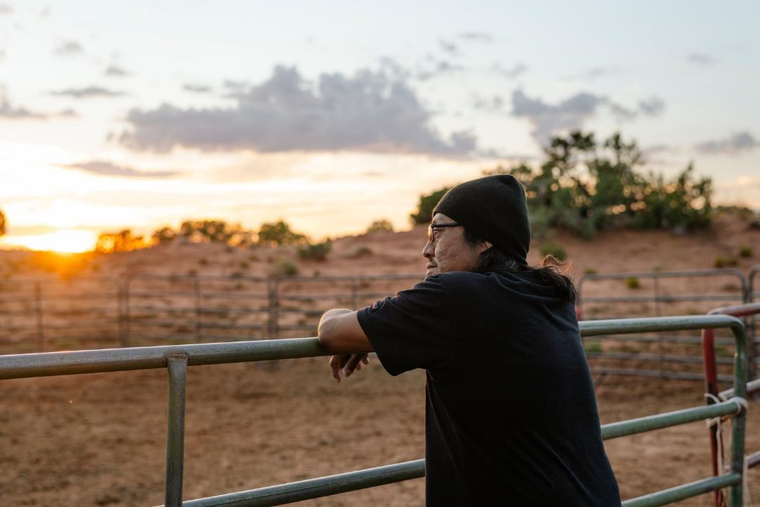 Nobert, a DaVita home dialysis patient, leans against a metal farm fence while he looks out at a sunset on Navajo Nation.