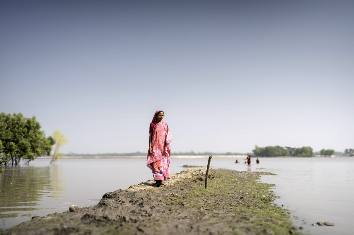 A woman stands on a narrow embankment surrounded by floodwater, with trees and people wading in the distance.