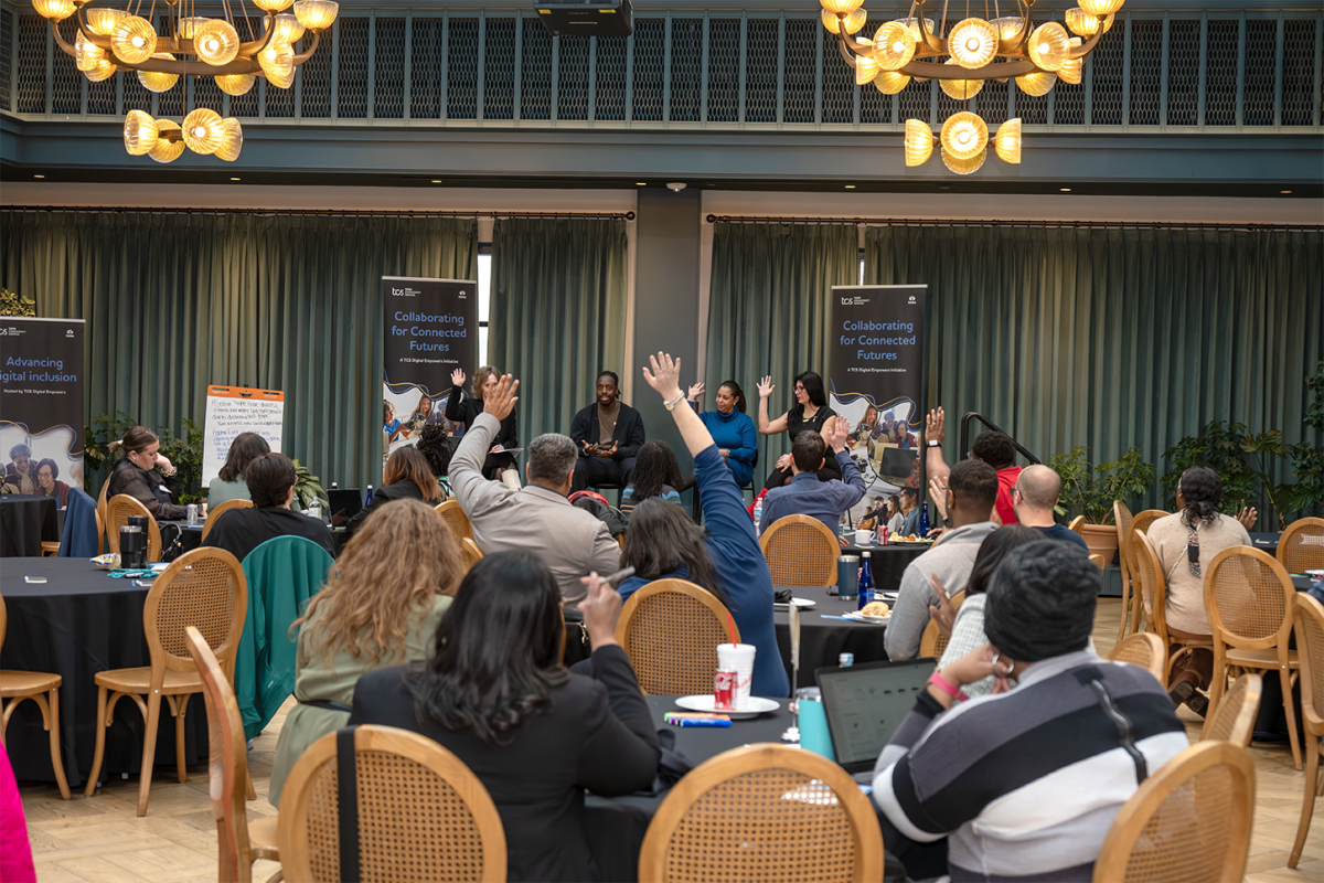 seated people raising hands at an event with speakers seated on stage
