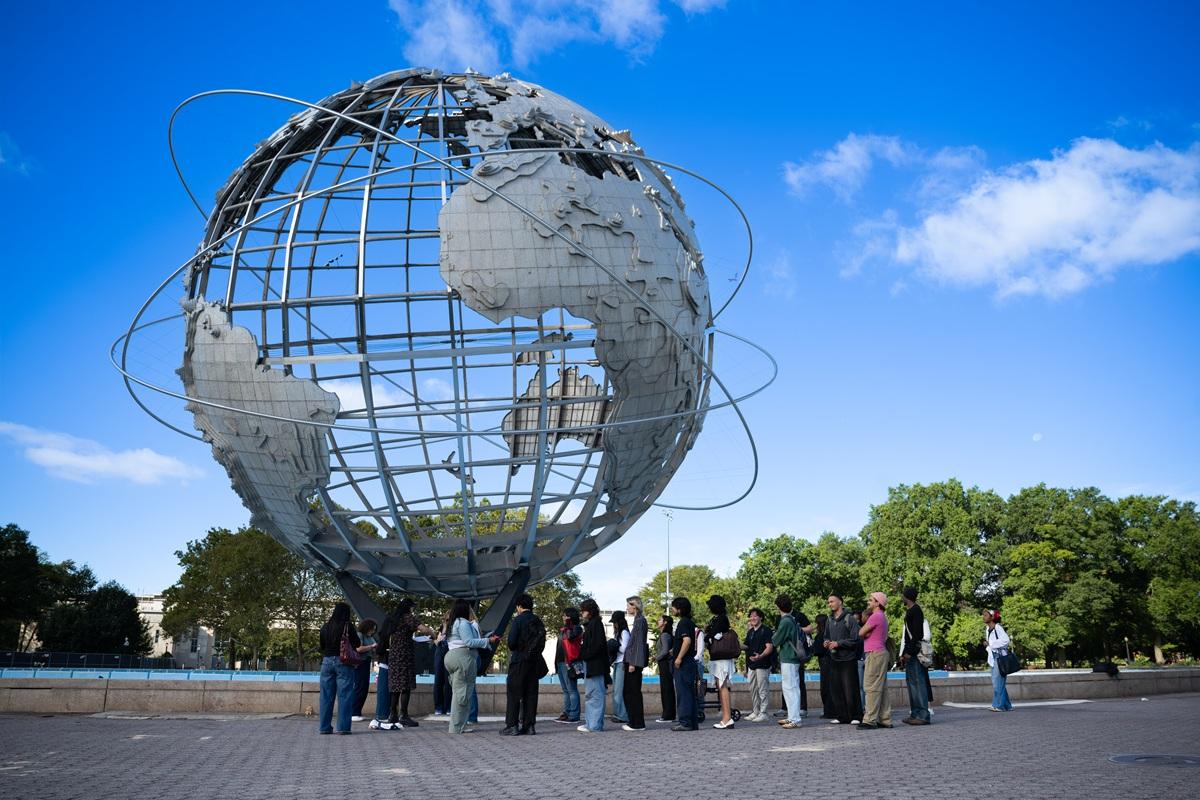 Group gathers outdoors beneath the Unisphere at Flushing Meadows Corona Park under a bright blue sky.