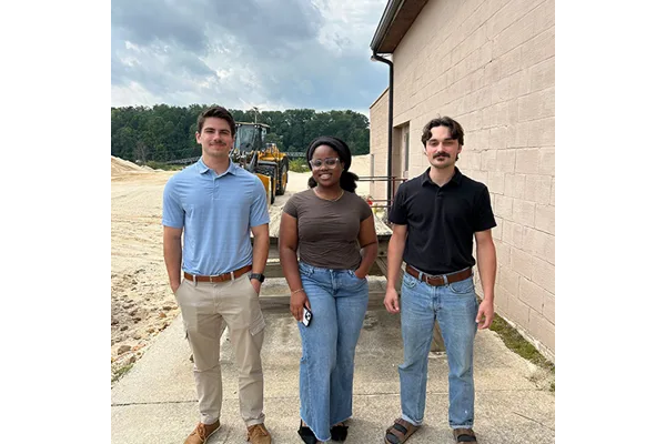 The three trainees standing together next to a building, with a tractor and trees in the background