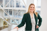 Maureen McHugh smiles at the camera and stands holding a railing at the top of a set of white steps at Bath & Body Works’ home office in Columbus, Ohio. Sun shines through a wall of windows in the background.