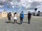 four people walking away from the camera on a gravel path. A row of Small brick buildings on their left and an open trailer on their right