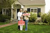 Young family of four in front of their new home holding a "sold" sign.