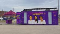 Exterior view of the Curiosity Cube™ mobile science lab featuring the message “Breakthroughs Begin with Curiosity” and an image of three student scientists.